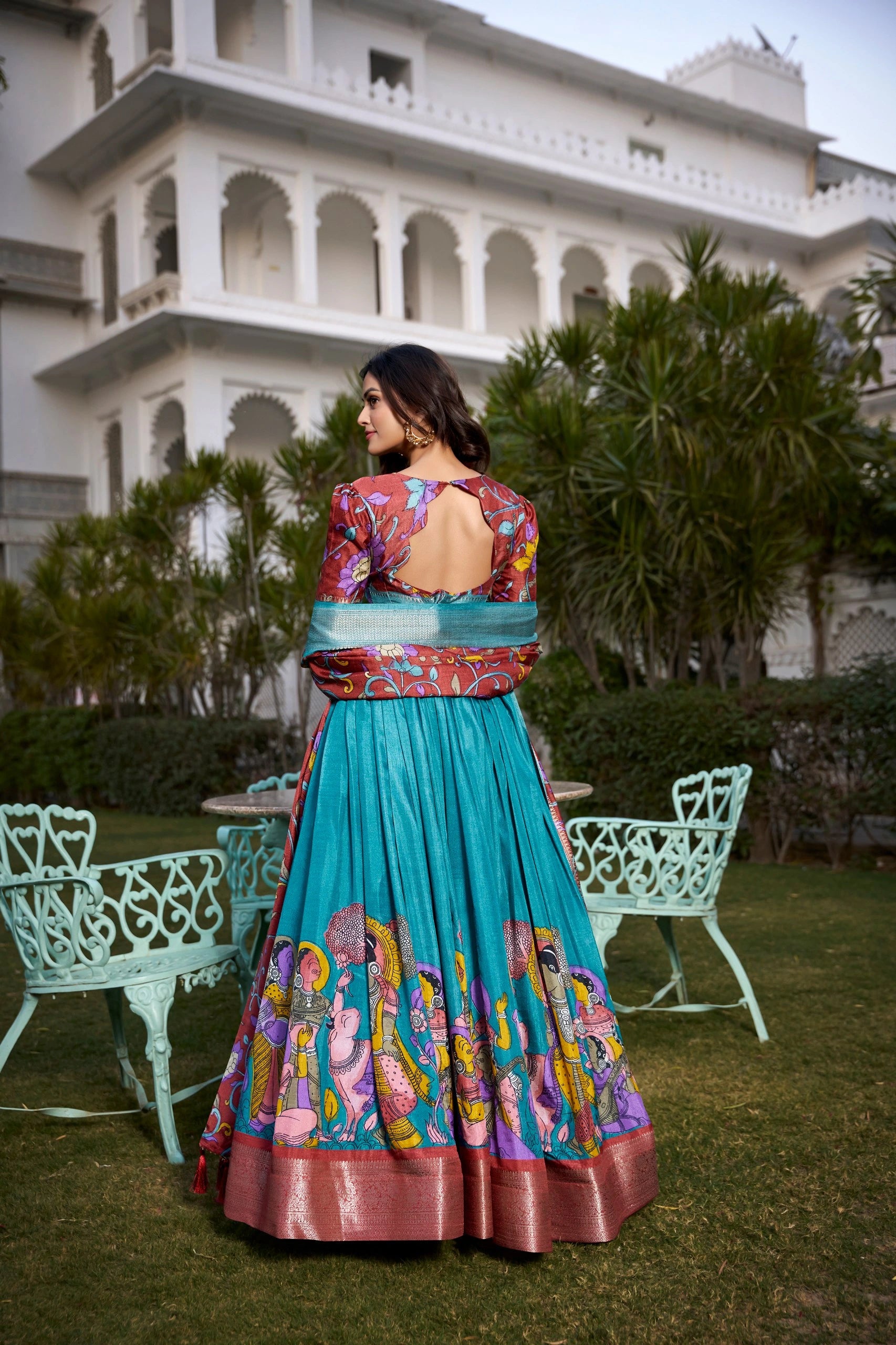 Woman in a colorful traditional outfit standing in front of a white building with greenery.